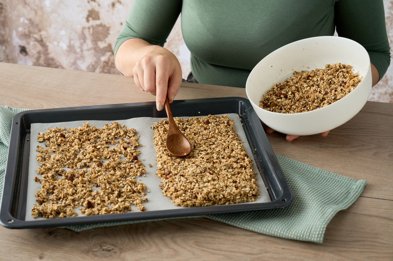 Person verteilt eine Granola-Mischung mit einem Holzlöffel auf einem mit Backpapier belegten Backblech und hält dabei eine weiße Schüssel.