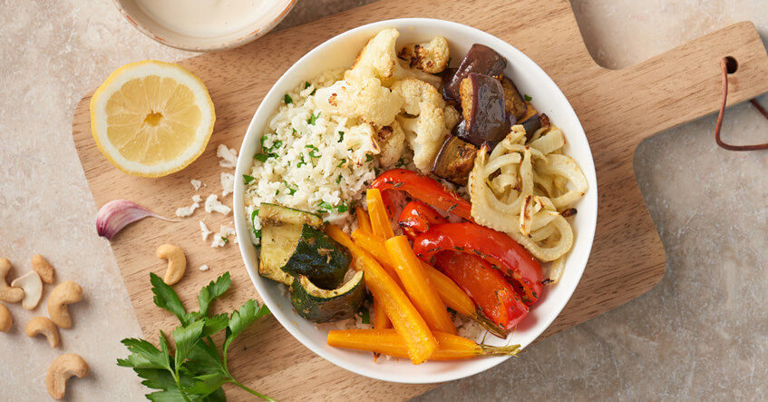 Eine Bowl mit gebackenem Ofengemüse, Reis und frischen Kräutern, angerichtet auf einem Holzbrett, daneben eine halbe Zitrone, Cashewkerne und Petersilie.