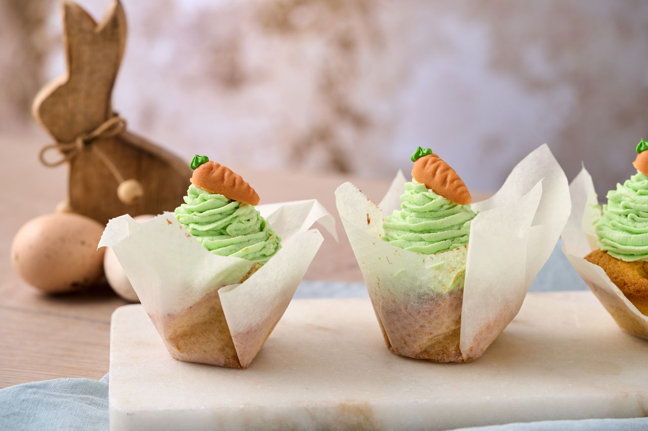 Karotten-Muffins mit grüner Creme und Marzipan-Möhre in weißem Backpapier, im Hintergrund ein Holz-Osterhase und Deko-Eier 