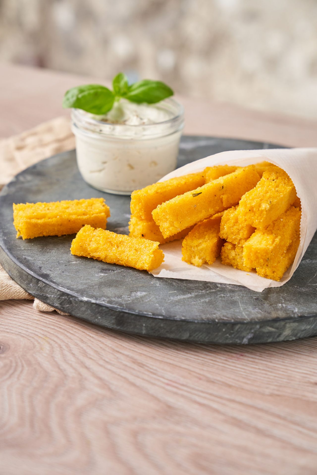 Knusprige Polenta-Pommes in weißer Backpapier-Tüte auf einem Teller, dazu ein Glas mit Dip und frischem Basilikum 
