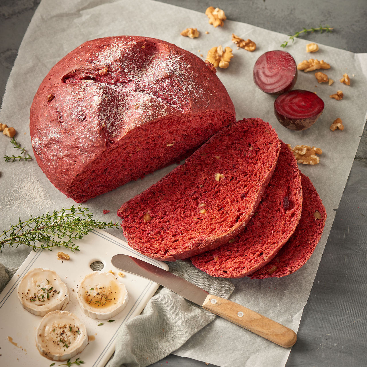 Rote-Bete-Brot mit Walnüssen, aufgeschnitten auf weißem Backpapier, daneben frische Rote Bete, Walnüsse, Thymian und eine kleine Platte mit Ziegenkäse und Messer