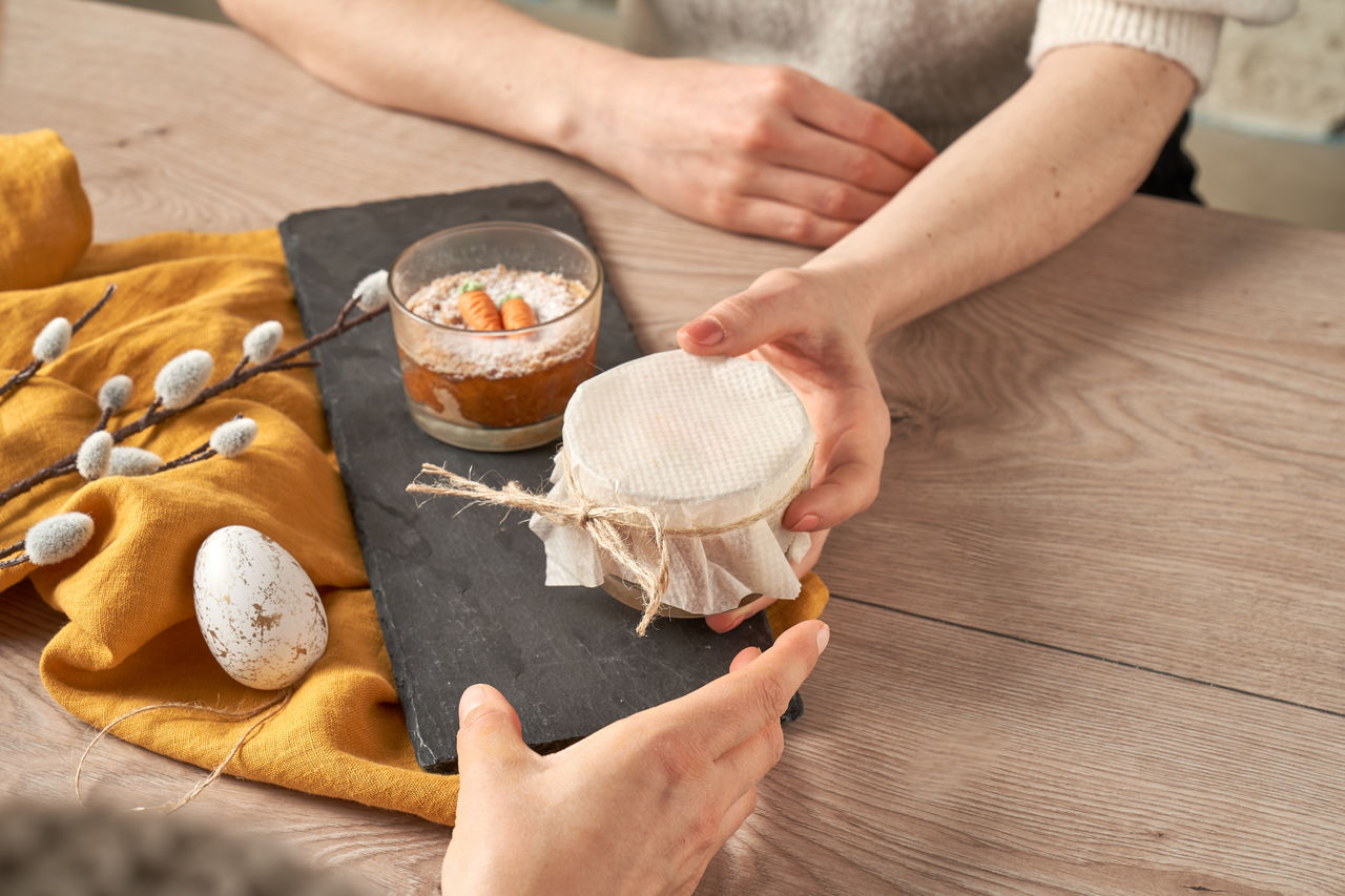 "Person übergibt ein Glas mit Karottenkuchen, das mit Toppits weißem Backpapier und Kordel abgedeckt ist. Daneben steht ein weiteres Glas mit Karottenkuchen auf einer Schieferplatte, dekoriert mit Frühlingszweigen und einem Ei auf einem gelben Tuch 
