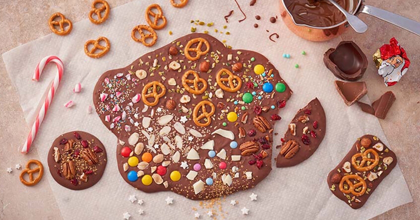 Geschmolzene Schokolade mit bunten Toppings wie Nüssen, Brezeln und Streuseln auf weißem Backpapier.