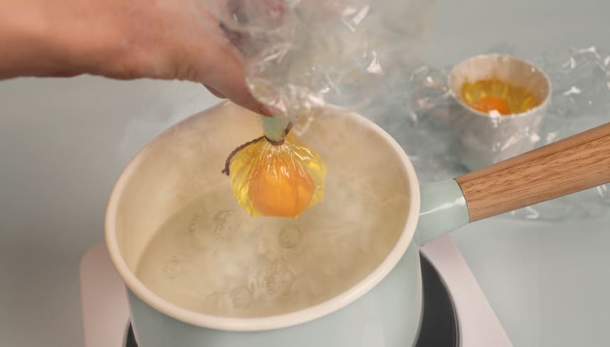 Eine Hand hält ein rohes Ei, das in Toppits Frischhaltefolie eingewickelt ist, über einen Topf mit kochendem Wasser. Im Hintergrund steht ein weiteres Ei in einem mit Folie ausgelegten Becher 