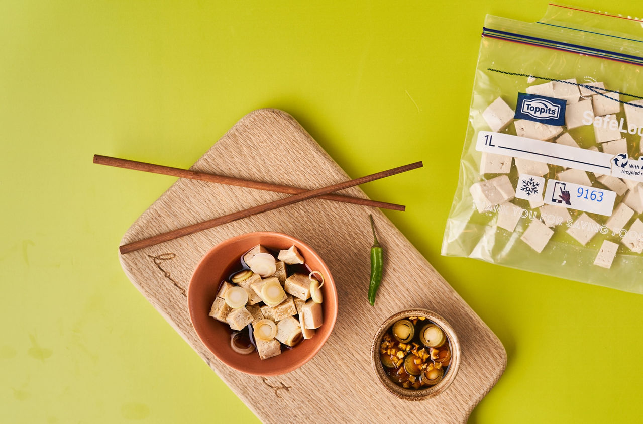 Toppits Gefrierbeutel mit Tofuwürfeln liegt neben asiatischem Gericht auf grünem Untergrund.