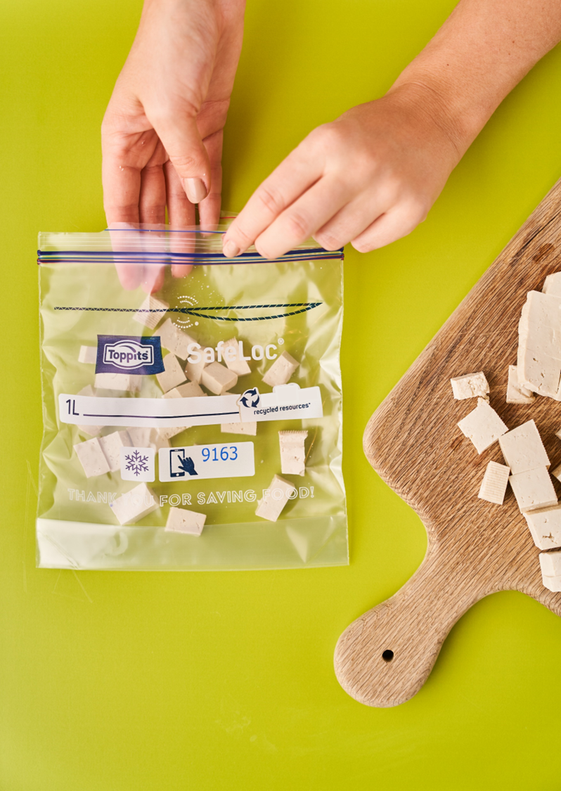 Zwei Hände verschließen einen Toppits SafeLoc Beutel mit gewürfeltem Tofu, daneben ein Holzbrett mit weiteren Tofuwürfeln auf grünem Untergrund 