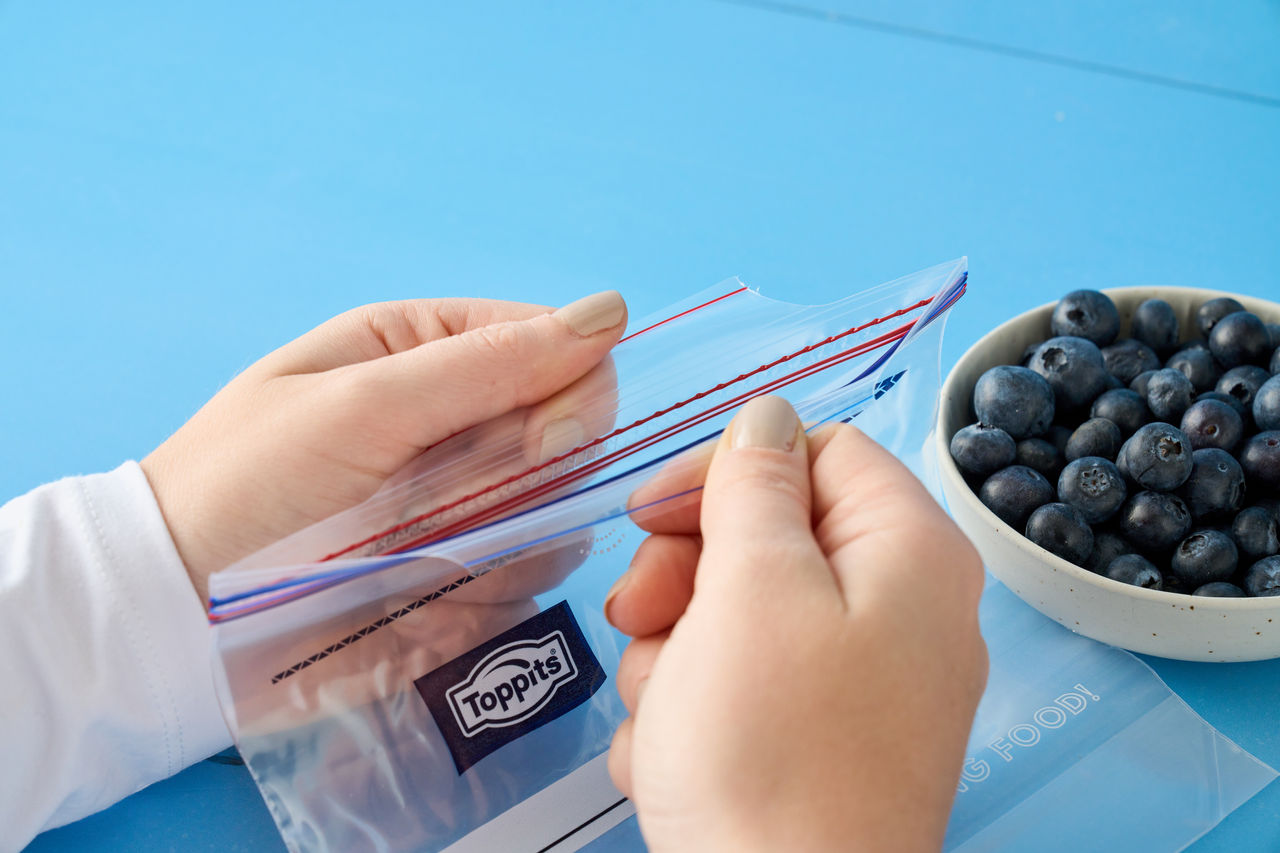 Zwei Hände öffnen einen leeren Toppits SafeLoc Gefrierbeutel, daneben steht eine Schale mit frischen Heidelbeeren auf einem blauen Untergrund