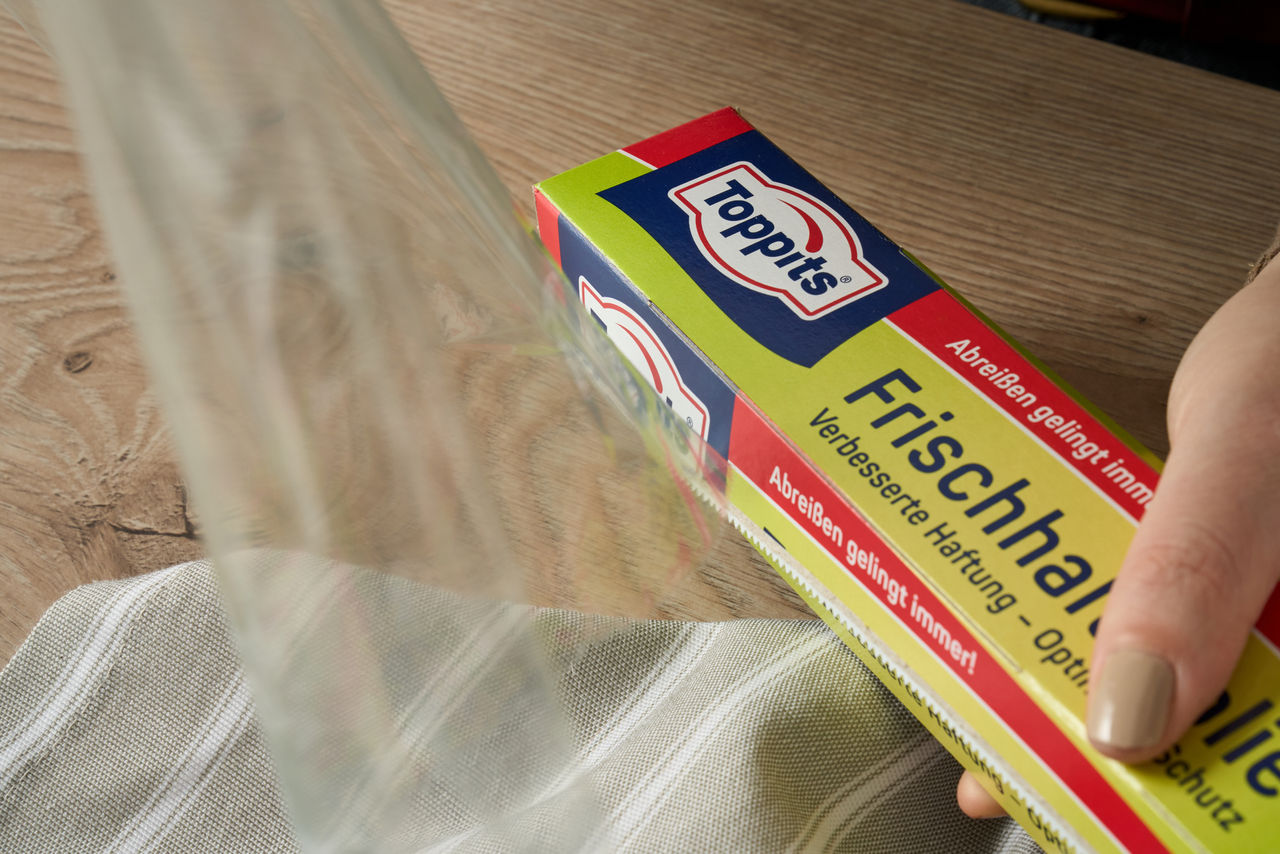 Person zieht Toppits Frischhaltefolie aus der Verpackung über eine gestreifte Tischdecke.