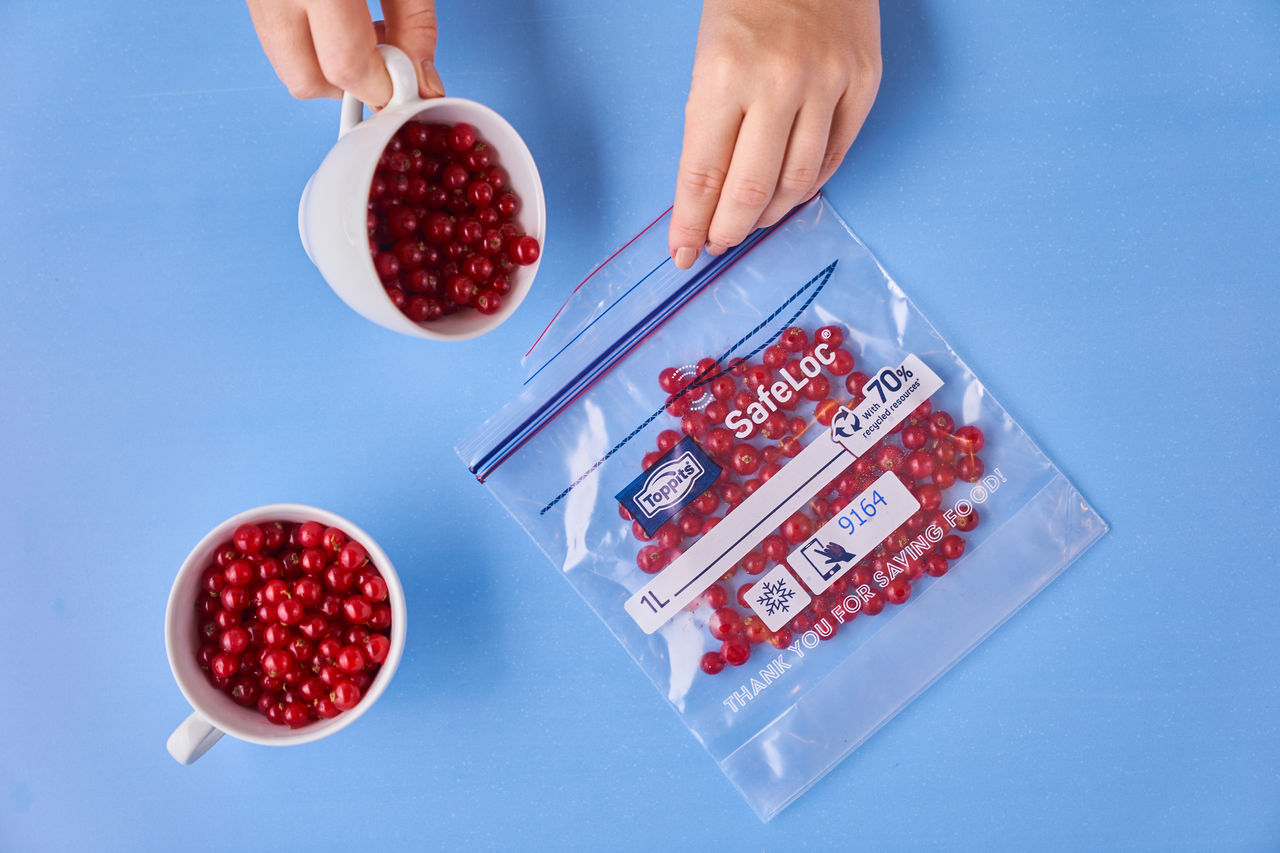 Zwei Hände füllen rote Johannisbeeren aus einer Tasse in einen durchsichtigen Toppits SafeLoc Gefrierbeutel auf einem hellblauen Untergrund. Neben dem Beutel steht eine weitere Tasse mit Johannisbeeren